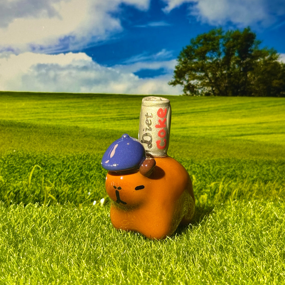 An orange pig-shaped figurine wearing a blue cap, standing in a grassy field with a Diet Coke can balanced on its back, against a backdrop of green hills and a blue sky.